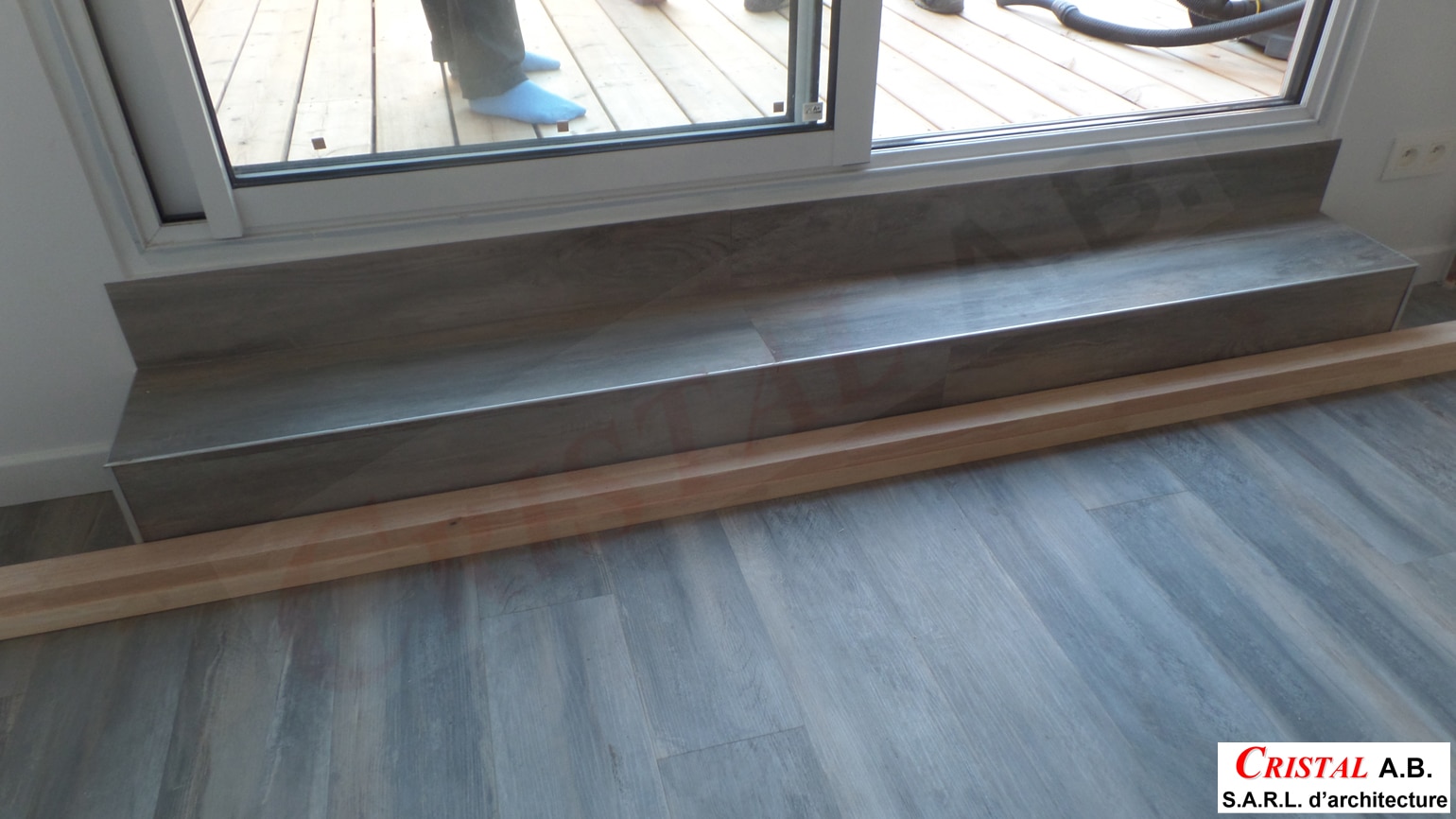 Escalier intérieur avec revêtement imitation bois gris devant une baie vitrée, logo Cristal A.B. S.A.R.L. d’architecture.