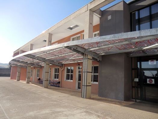 Façade d'école moderne avec un grand préau métallique à motifs rouges sur une cour ensoleillée.