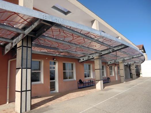 Auvent métallique courbé à motifs rouges devant un bâtiment scolaire moderne aux murs saumon.