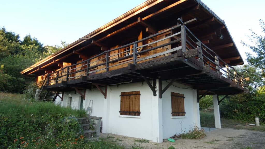 Chalet de montagne en bois sur base blanche avec balcon et volets, entouré de verdure sous un ciel clair.