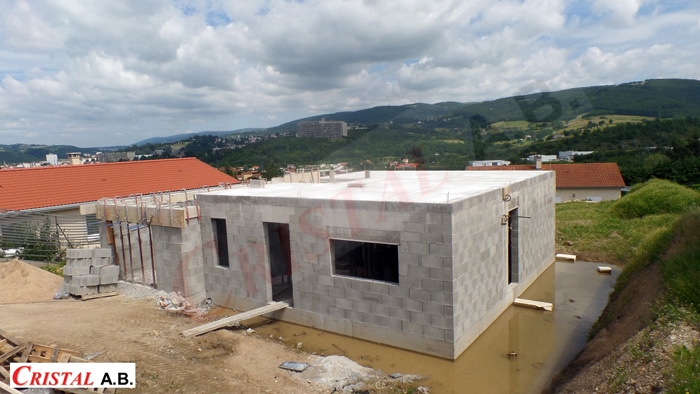Maison en construction Cristal A.B. en blocs de béton avec toit plat entourée d'eau devant un paysage de montagne.