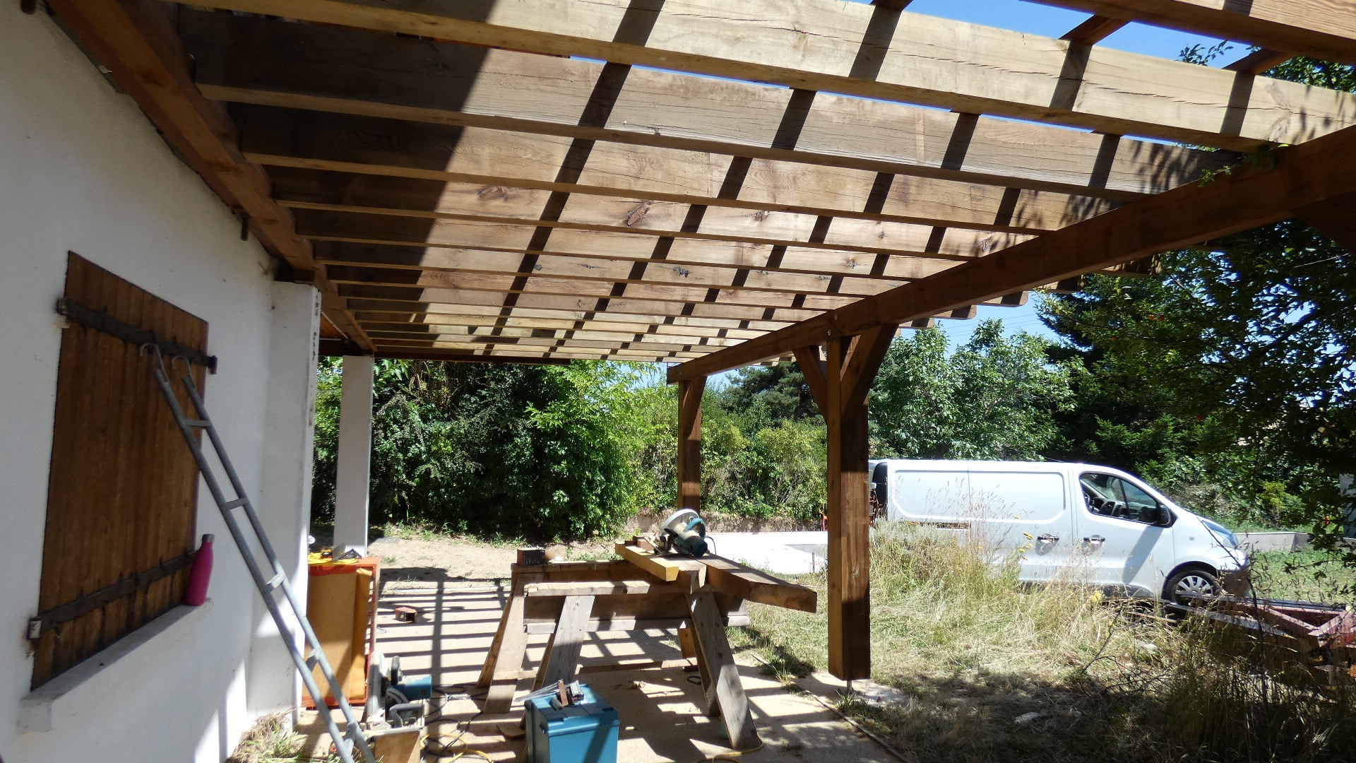 Construction d'une pergola en bois adossée à une maison avec outils de menuiserie et charpente apparente.