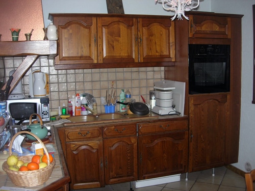 Cuisine rustique avec meubles en bois brun, four encastré, micro-ondes et panier de fruits sur plan de travail carrelé.