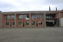 Bâtiment scolaire rose avec piliers en béton, grandes fenêtres et cour d'école goudronnée.