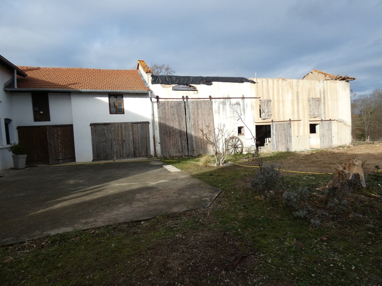 Grange ancienne en pierre avec toit de tuiles, portes en bois et roue de charrette devant une cour bétonnée.