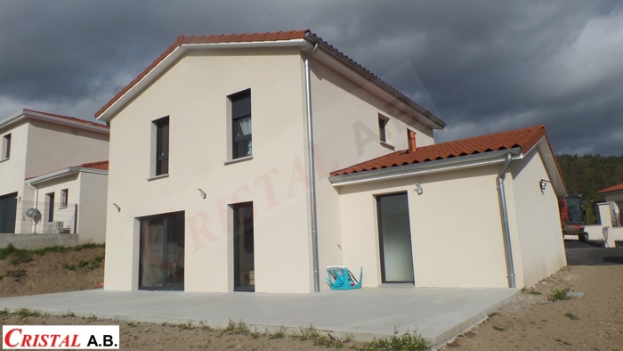 Maison moderne à façade beige, toiture en tuiles rouges et terrasse béton. Logo CRISTAL A.B. en bas à gauche.
