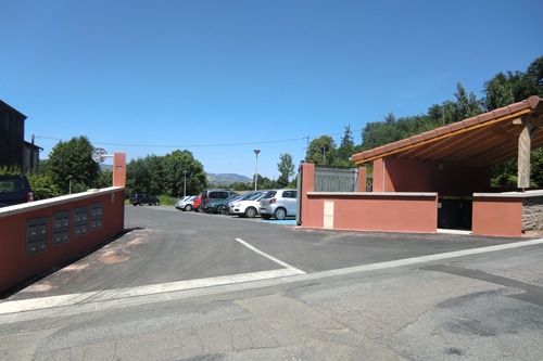 Parking avec voitures, boîtes aux lettres sur mur ocre et abri couvert sous un ciel bleu dégagé.