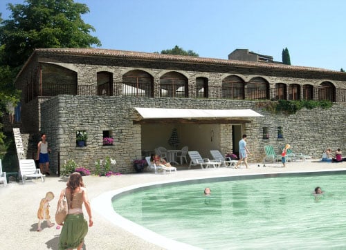 Personnes se détendant au bord d'une piscine devant une villa en pierre avec terrasse et arches sous un ciel bleu.