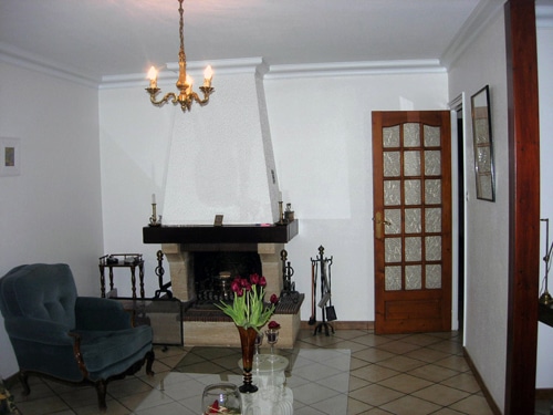 Salon classique avec cheminée, fauteuil velours bleu, lustre élégant et fleurs sur table basse vitrée.