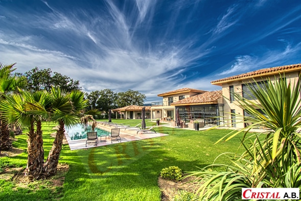 Villa de luxe avec piscine et jardin Cristal A.B., palmiers et pelouse sous un ciel bleu avec nuages filandreux.