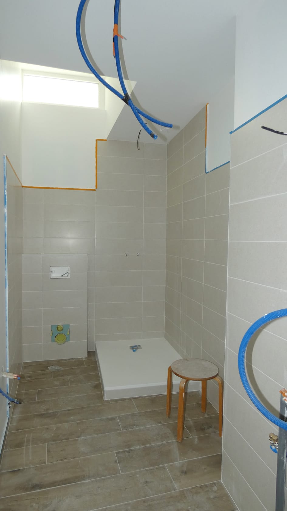 Rénovation de salle de bain : carrelage beige, sol imitation bois, receveur de douche et conduits de plomberie.