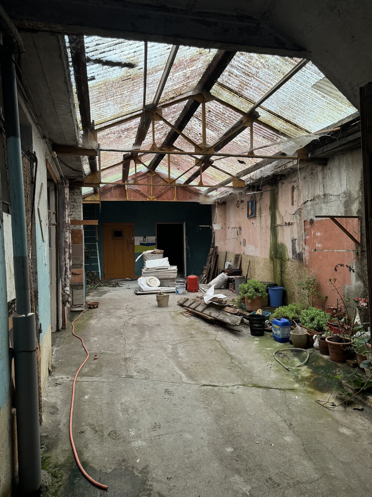 Cour intérieure délabrée avec toit en tôle translucide, vieux outils et plantes en pots sur sol en béton.