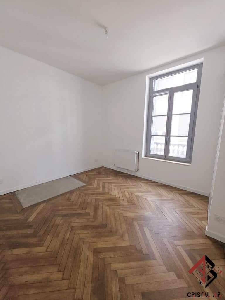 Salon vide avec parquet en chevrons, murs blancs et fenêtre grise.