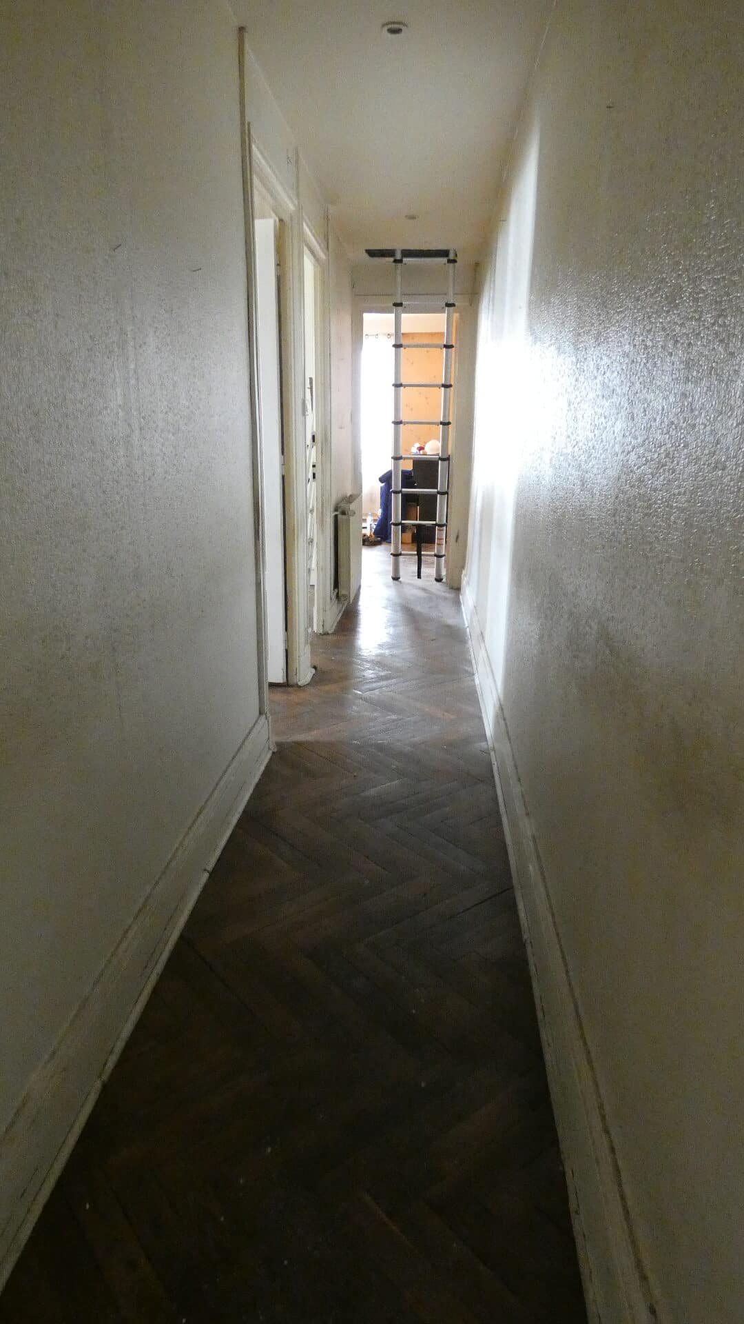 Couloir étroit avec parquet ancien en chevrons, murs blancs défraîchis et une échelle télescopique au bout.