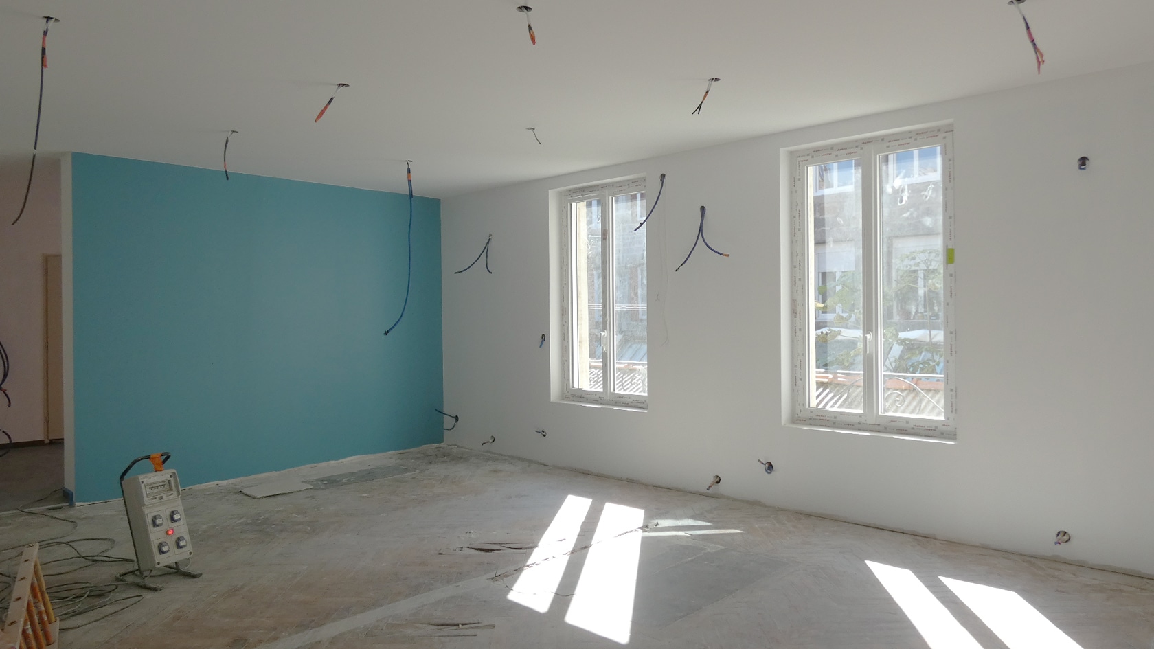 Pièce en rénovation avec mur bleu canard, câbles électriques au plafond et lumière naturelle sur sol brut.