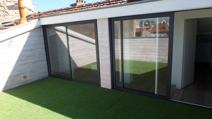 Terrasse de toit moderne avec gazon synthétique et larges baies vitrées coulissantes ouvrant sur l'intérieur.
