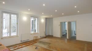 Salon lumineux en travaux avec parquet chevron, grandes fenêtres et spots encastrés au plafond.